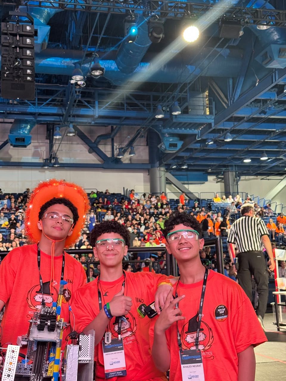 Three team members in orange team shirts and safety glasses smiling inside a large competition arena.