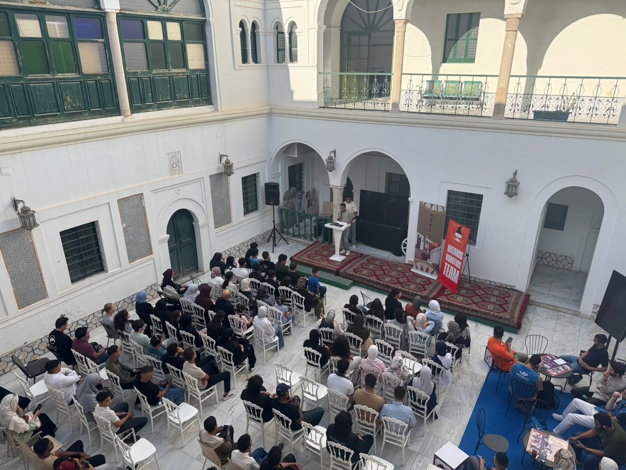 Seen from above: an audience seated in a white courtyard listens to a speaker, next to a Wizards Robotics Team banner.